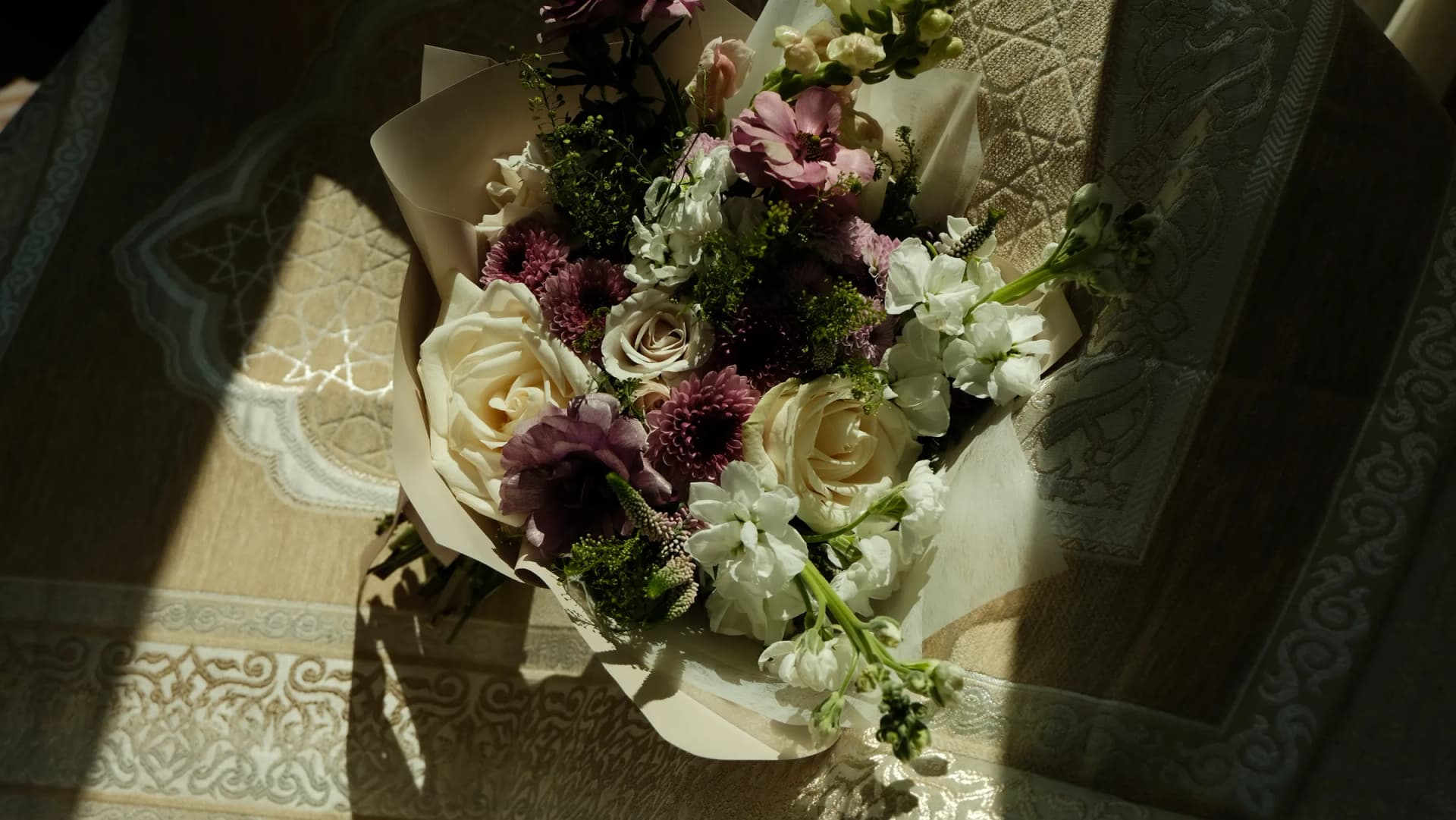 A curated bouquet of cream roses, dusty mauve chrysanthemums, and white stock wrapped in cream paper, bathed in warm golden-hour light